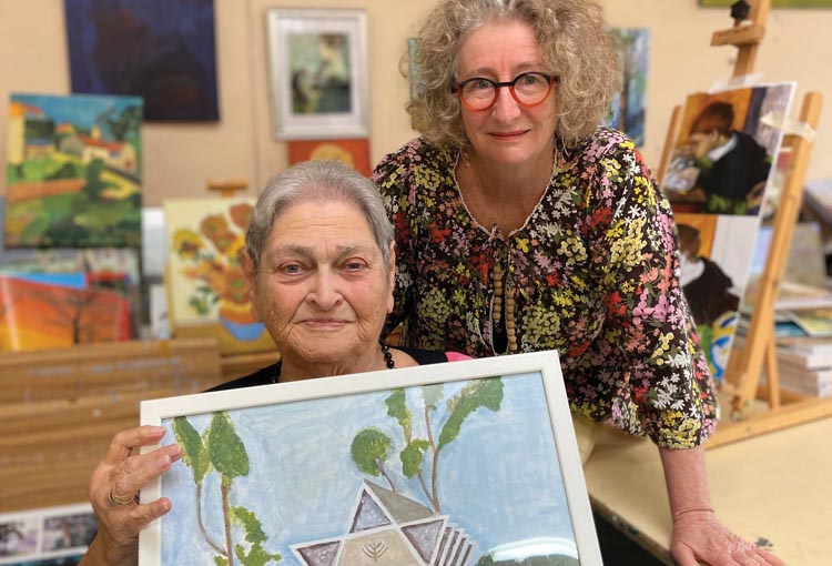 Senior woman Bertha Podobransky sitting down and holding her painting of the former Bankstown Synagogue. Standing next to her is Hunters Hill Art Therapist Lea Dalgleish.